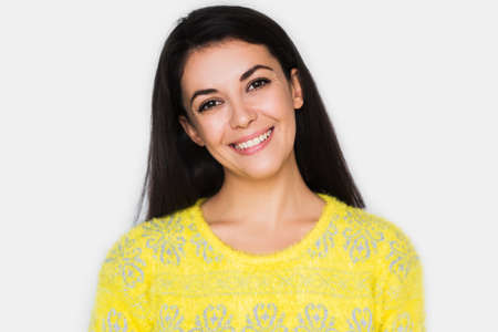 Portrait of brunette cheerful woman with healthy toothy smile, look in front with hands on hair. Young happy pretty woman wearing yellow sweater on light grey background. Positive and happiness mood.の写真素材
