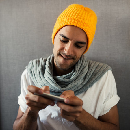Attractive young man playing the game on device, telephone. Wearing orange hat and gray scarf, on...