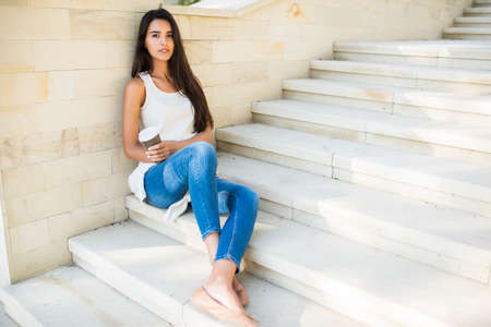 Full body portrait of beautiful brunette woman sitting on the steps of a park, on lake background. Smiling with a cup of coffee or tea. Looking at the camera.の写真素材