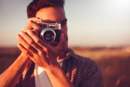 Close-up beautiful portrait of young handsome man with vintage camera, take a picture, on field background, and on sunset. Autumn, summer, mood. Photographyの写真素材
