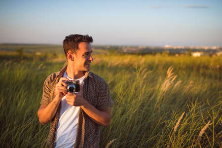 Handsome young traveler man with vintage camera,on a green meadow background. Travel mood. Photography. Relaxation on a field and sunset. Explore nature.の写真素材