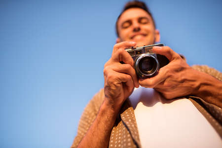 Bottom view of smiling handsome young man with vintage camera, take a picture on sky background. Autumn. Summer. Mood background. Photography.の写真素材