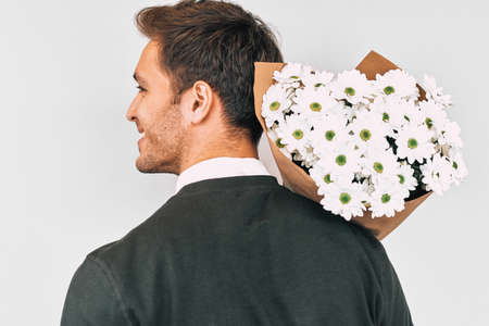 Rear view shot of happy handsome man smiling holding a bouquet of white flowers. Attractive male with a bunch of flowers preparing for a date with girlfriend. Valentine's Day. Mother's Day conceptの写真素材