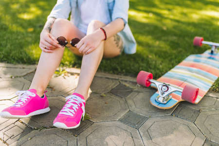 Beautiful legs of woman wearing pink shoes  with sunglasses in hand sitting in park on green grass background in a blue shirt. Sporty female relaxing after longboarding outdoor.の写真素材