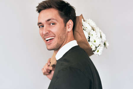 Handsome young man smiling and delivering a bouquet of white flowers. Attractive male model with a bunch of flowers preparing for a date with girlfriend. Valentine's Day. Mother's Day conceptの写真素材