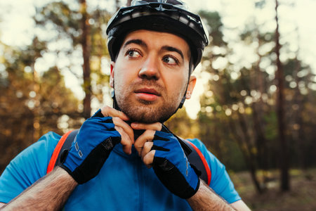 Close up portrait of serious and thoughtful cyclist close the protective helmet outdoors and looking away, thinking about the route. Travel concept. Sport lifestyle. Young rider wearing blue t-shirt.の写真素材