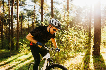 Handsome young European male wearing protective helmet and black sports clothing and glasses riding bicycle outdoors in forest or mountains, enjoy morning ride, having calm and confident expression.の写真素材