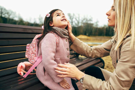 Candid side view of blonde pretty mother carring her cute girl kid sitting on a bench outdoors. Loving blonde young woman and her cute daughter spending time together in the city park. Mother's Dayの写真素材
