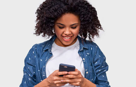 Indoor close up portrait of smiling African American young woman using cellphone, messaging to her friend. Happy dark skinned female chatting with her boyfriend, looking at screen of smart phone.の写真素材