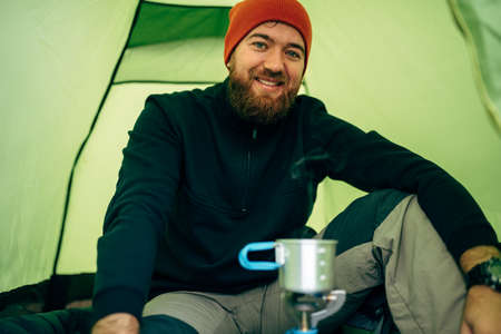 Portrait of happy young man explorer drinking hot beverage in mountains. Traveler bearded man wearing red hat, sitting near to camping tent, preparing a mug of tea after hiking. Travel, lifestyleの写真素材