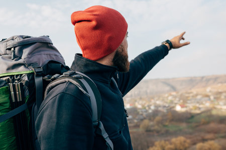 Rear view image of young hiker male wearing red hat, hiking in mountains, showing something to the horizon. Traveler bearded man relaxing after mountaineering. Travel, people and lifestyle conceptの写真素材