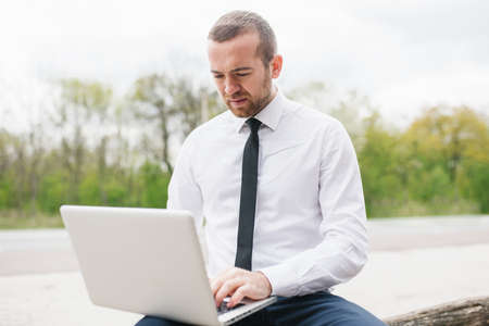 Fashion smart businessman wearing white shirt, typing on his computer making report of his firm using wireless. Handsome man working on laptop outdoor in the park. Technology and business concept.の写真素材