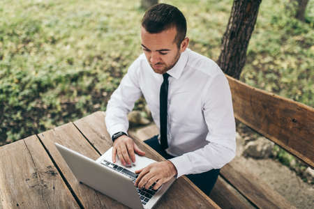 Top view of smart businessman wearing white shirt, typing on his computer making report of his firm. Handsome man working on laptop outdoor in the park. Technology, occupation and job concept.の写真素材