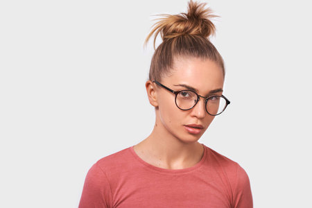 Attractive blonde young woman wears spectacles and casual pink blouse, looks seriously directly into...
