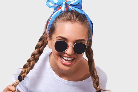 Close up portrait of young pretty woman wearing white t-shirt, blue headband and sunglasses with braids hairstyle, isolated on white backgroundの写真素材