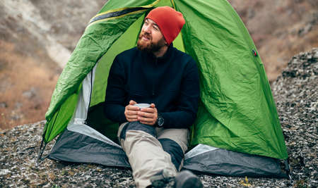 Horizontal outdoors image of traveler bearded male sitting near the camping tent, relaxing after mountaineering. Young hiker man enjoy the air in the mountain. Travel, people and healthy lifestyleの写真素材