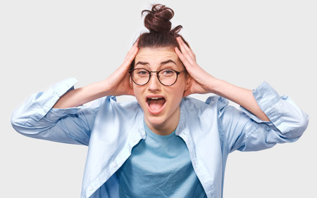 Studio portrait of unhappy young woman with hair bun, opens mouth widely, screams loud with hands on...