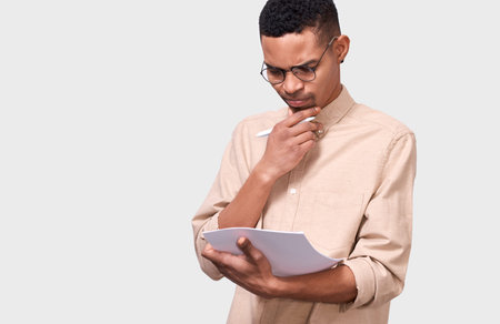 Serious young businessman in beige shirt analyzing financial reports posing on white studio...