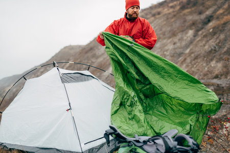 Horizontal outdoors image of authentic young hiker man prepares his tent in mountains. Traveler bearded male relaxing after mountaineering. Travel, people and healthy lifestyle conceptの写真素材