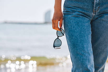 Cropped shot view of beautiful tanned woman hand with black sunglasses walking on the beach against calm sea, blue sky background with copy space for your content, summer vacation and travel conceptの写真素材