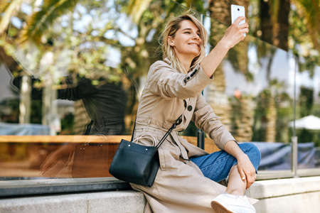 Side view of beautiful young woman smiling while holding smartphone and making self portrait, strolling outdoor in the city street against palms. Happy female taking selfie on mobile phone. Travelの写真素材