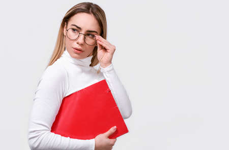 Serious young businesswoman wearing white blouse and round trendy eyeglasses with red folder in hand, with blank copy space for your text. European female office worker posing on white studio wall.の写真素材