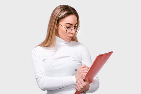 Serious young woman wearing white blouse and round transparent eyeglasses with red folder in hands,...