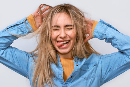 Close up studio portrait of funny young blonde woman with hands in the hair dressed casual blue...
