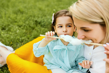 Closeup portrait of little girl eating cotton candy with her mom, sitting on the green grass in the...
