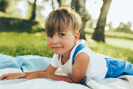 Cute little boy lying on the blanket and looking to the camera. Happy child enjoying summertime in the park. Handsome kid smiling and relaxing on sunlight outdoors. Childhood conceptの写真素材