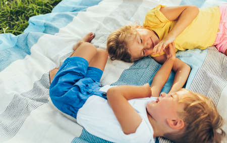 Horizontal image of playful children, sister and brother lying on the blanket, looking to each other. Happy little boy and little girl enjoying summertime in the park. Cheerful kids playing outdoorsの写真素材