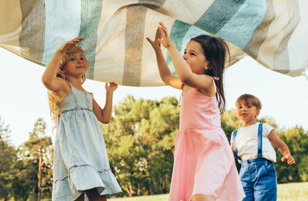 Children playing under the blanket, jumping and dancing together. Happy little boy and little girls enjoying summer day in the park. Cheerful kids playing outdoors. Childhood conceptの写真素材