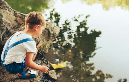 Rear view image of cute little boy launch paper ship on the lake in the park. Adorable kid boy playing with a boat. Curious child sailing a toy boat by the waters' edge outdoors. Childhood and ecologyの写真素材