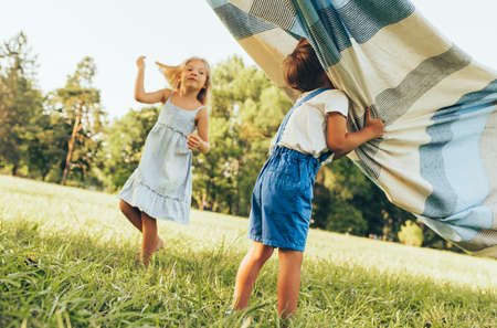 Outdoors image of children playing under the blanket, jumping and dancing together. Happy little boy and little girl enjoying summer day in the park. Cheerful kids playing outdoors. Childhood conceptの写真素材