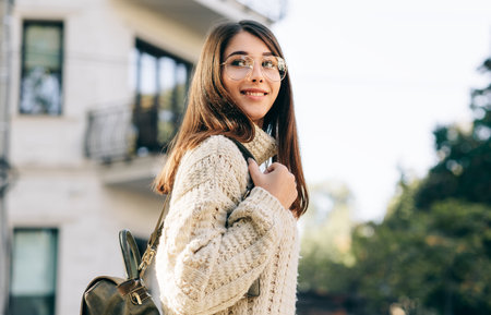 Smiling pretty girl student looking back, standing on the street in the morning, wearing knitted...