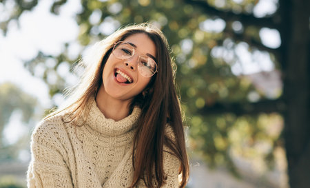 Close-up outdoor portrait of funny student woman showing tongue out, looking to the camera, wearing...