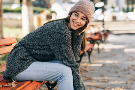 Image of pretty smiling student female looking to the camera, sitting on the bench in the city...
