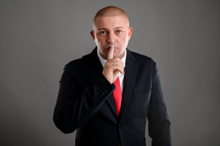 Portrait of cool businessman wearing elegant black suit and red tie placing fingers on lips as if to...
