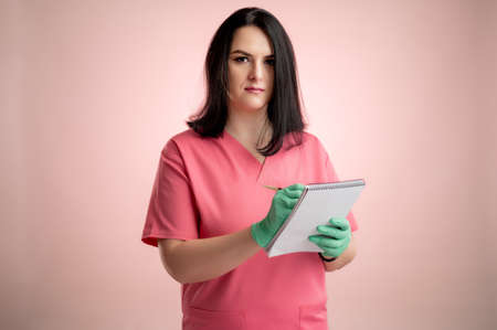Portrait of beautiful woman doctor with stethoscope wearing pink scrubs, with brown hair, takes notes, looking at the camera posing on a pink isolated backround.の写真素材