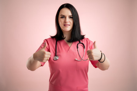 Portrait of beautiful woman doctor with stethoscope wearing pink scrubs, showing thumbs up with both hands posing on a pink isolated backround.の写真素材