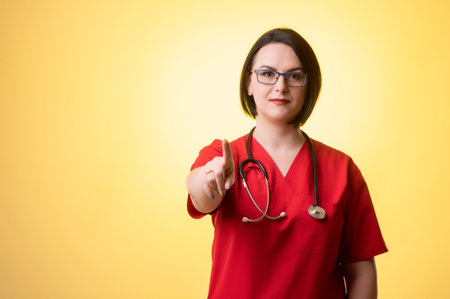 Portrait of beautiful woman doctor with stethoscope, wearing red scrubs, pointing her finger posing on a yellow isolated background.の写真素材
