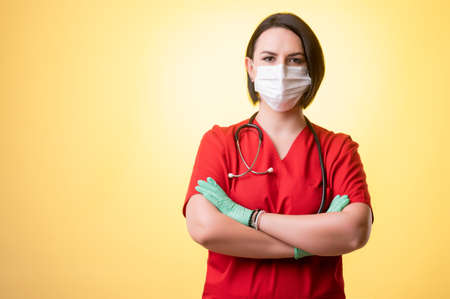 Portrait of beautiful woman doctor with stethoscope wearing red scrubs, with protective mask posing on a yellow isolated background.の写真素材