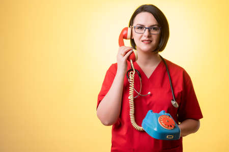Portrait of beautiful woman doctor with stethoscope wearing red scrubs, with brown hair, displeased face and gestures and talking on old phone looking at camera, posing on a yellow isolated background.の写真素材