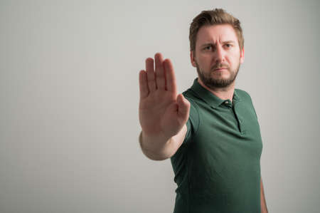 Portrait of serious stylish attractive man with thick beard, dressed in casual green t shirt showing stop gesture isolated on gray background with copy space advertising areaの写真素材