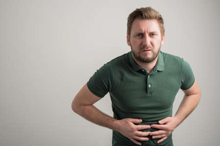 Portrait of serious stylish attractive man with thick beard, dressed in casual green t shirt gesturing stomach ache isolated on gray background with copy space advertising areaの写真素材