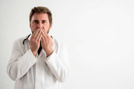 Portrait of male doctor with stethoscope in medical uniform covering mouth like mute concept, posing on a white isolated backgroundの写真素材