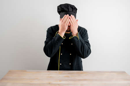 Portrait of young male chef in black uniform covering eye like blind concept posing on a white isolated backgroundの写真素材