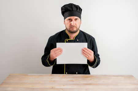 Portrait of young male chef in black uniform holding a white paper, upset posing on a white isolated background.の写真素材