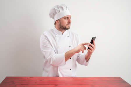 Portrait of young male chef in white uniform with blonde hair, holding smartphone, texting messages posing on a white isolated backgroundの写真素材