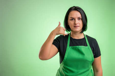 Female cook with green apron and black t-shirt, showing call me gesture isolated on green backgroundの写真素材